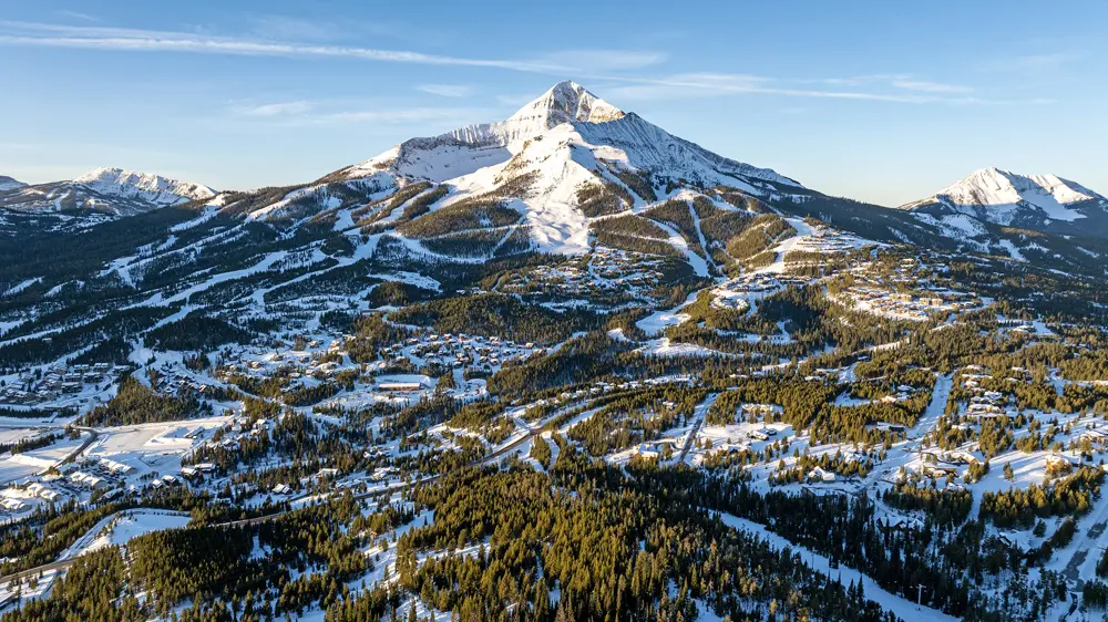 Snowy mountain resort with forested slopes, ski runs, and buildings beneath a blue sky.