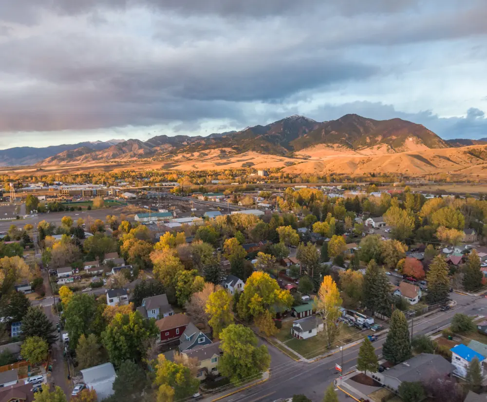 Aerial view of a town and autumn trees beneath sunlit foothills and mountains.